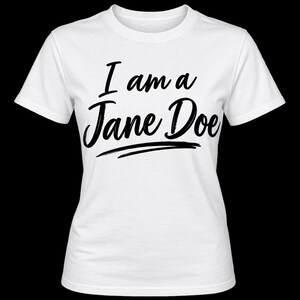 Op de afbeelding: Een wit t-shirt met korte mouwen en de zwarte tekst "I am a Jane Doe" in een gestileerd scriptlettertype op de borst. Het ontwerp bevat een horizontale zwarte penseelstreek onder de tekst, gecentreerd op het t-shirt.