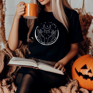 Könnte beinhalten: Schwarzes T-Shirt mit einem weißen Grafikdesign, das einen Halbmond, Sterne und Hände zeigt. Der Text "DO NO HARM TAKE NO SHIT" ist um das Design herum gedruckt. Die Person hält einen orangefarbenen Becher und ein Buch, mit einem Kürbis in der Nähe.