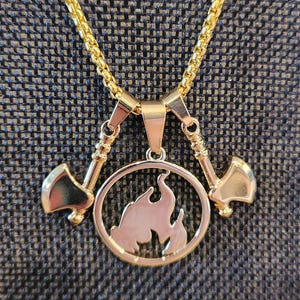 May include: Gold-coloured necklace featuring a pendant with a flame design inside a circle, flanked by two small axes. The chain has a textured, woven appearance. The necklace is displayed against a dark, textured background.