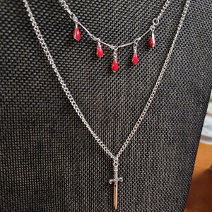 May include: A silver chain necklace with a small silver sword pendant. A second chain with red teardrop-shaped charms hangs above the sword pendant.