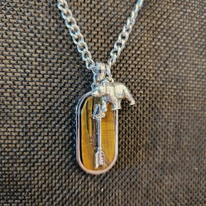 May include: Silver-toned necklace with a pendant featuring a tiger's eye stone. The pendant has a silver bear and arrow design. The chain is a curb link style, and the background is a textured brown.