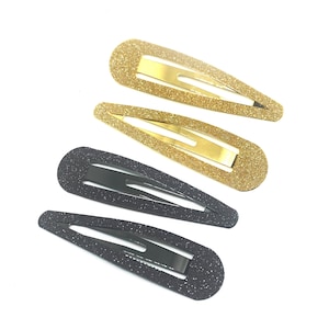以下が含まれることがあります： 金と黒のグリッターヘアクリップ4個。2つのゴールドクリップはゴールドの金属製の留め金、2つのブラッククリップはブラックの金属製の留め金が付いています。クリップは楕円形で、白い背景にあります。