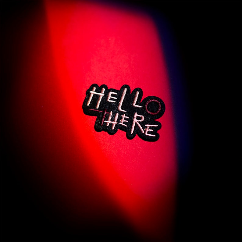 May include: A black embroidered patch with the words "HELLO THERE" in white, with a red line through the word "THERE". The patch is on a red surface with a black background.