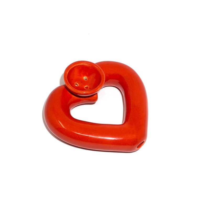 May include: A red ceramic heart-shaped pipe with a bowl on top. The pipe is designed with a hole in the center of the heart.