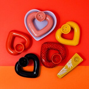 May include: Six heart-shaped ceramic pipes in various colors, including pink, yellow, black, and red. The pipes have a hole in the center and a small hole on the side.