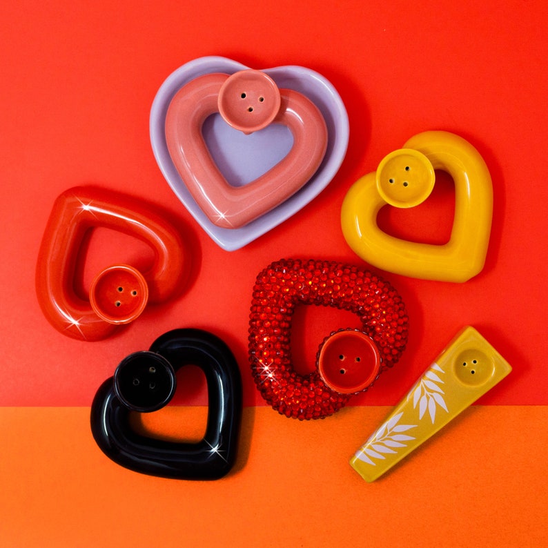 May include: Six heart-shaped ceramic pipes in various colors, including pink, yellow, black, and red. The pipes have a hole in the center and a small hole on the side.