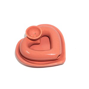 May include: A pink ceramic heart-shaped incense burner with a small bowl on top. The bowl has three holes in it.