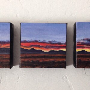May include: Three small paintings of a desert landscape with a sunset sky. The sky is a gradient of blue, purple, orange, and yellow. The mountains are silhouetted against the sky. The foreground is a dark brown.