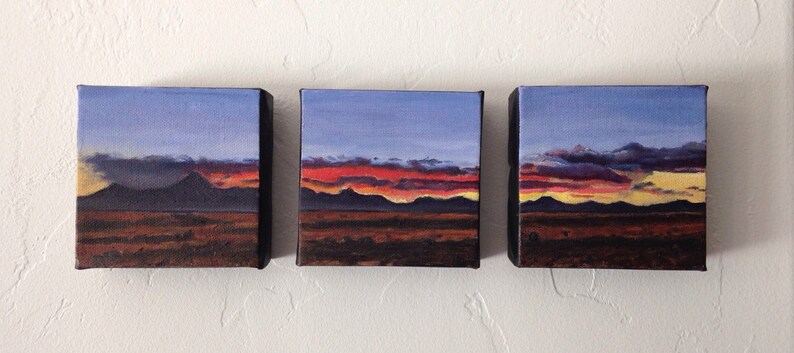 May include: Three small paintings of a desert landscape with a sunset sky. The sky is a gradient of blue, purple, orange, and yellow. The mountains are silhouetted against the sky. The foreground is a dark brown.