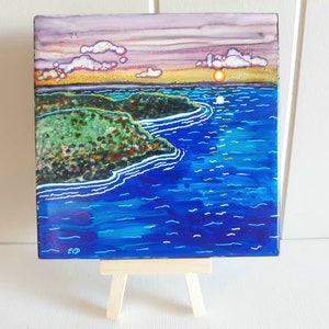 May include: A small painting on a wooden easel depicts a seascape with a green island in the foreground and a setting sun in the background. The sky is a gradient of purple, pink, and orange. The water is a deep blue with white lines representing waves.