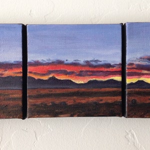 May include: A triptych painting depicting a sunset over a mountain range. The sky is a gradient of blue, purple, and orange, with clouds in the upper portion of the painting. The mountains are silhouetted against the sunset, and the foreground is a dark brown field.