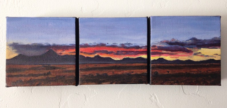 May include: A triptych painting depicting a sunset over a mountain range. The sky is a gradient of blue, purple, and orange, with clouds in the upper portion of the painting. The mountains are silhouetted against the sunset, and the foreground is a dark brown field.