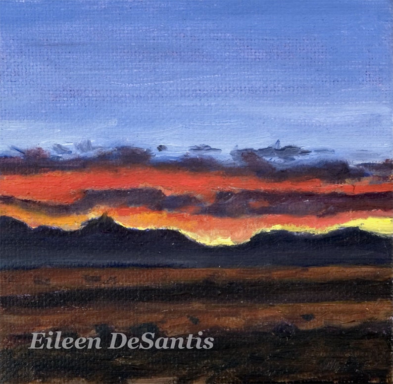 May include: A painting of a sunset over a mountain range. The sky is a deep blue with streaks of orange, red, and yellow. The mountains are silhouetted against the sky. The artist's signature, Eileen DeSantis, is in the bottom right corner.