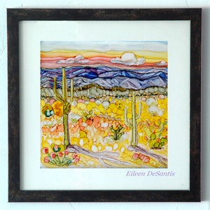 May include: A framed painting of a desert landscape with a sunset sky, purple mountains, and yellow and green cacti. The artist's signature, Eileen DeSantis, is in the bottom right corner.