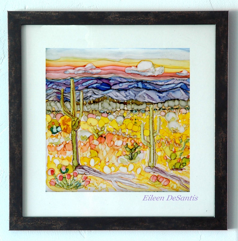 May include: A framed painting of a desert landscape with a sunset sky, purple mountains, and yellow and green cacti. The artist's signature, Eileen DeSantis, is in the bottom right corner.