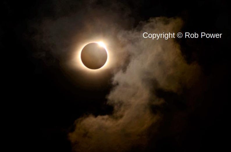 Pu&ograve; includere: Un'eclissi solare totale con la corona del sole visibile come un anello luminoso attorno alla luna, parzialmente oscurata da nuvole. Copyright &copy; Rob Power