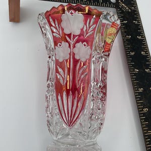 May include: A clear crystal vase with a red interior and white floral design. The vase has a flared top and a detailed, textured pattern. A small sticker is visible on the side. The vase is approximately 15 cm tall.