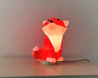 May include: A red geometric fox-shaped lamp with a white interior. The lamp is lit and casts a warm glow. The lamp is sitting on a wooden base.