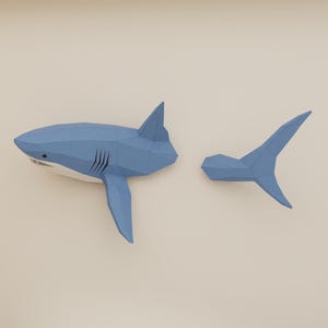 Könnte beinhalten: Eine hellblaue, geometrische Papierhai-Skulptur mit abgetrenntem Schwanz. Der Hai hat einen weißen Bauch, schwarze Kiemen und ein kleines Auge. Der Schwanz ist ebenfalls hellblau und hat eine spitze Form. Die Skulptur wird vor einem neutralen Hintergrund gezeigt.