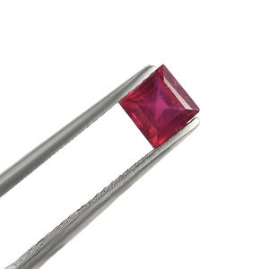 May include: A square-shaped, red ruby gemstone held by tweezers.