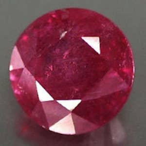 May include: A deep red ruby gemstone with a faceted cut. The gemstone is round and has a high polish.
