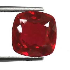 May include: A deep red gemstone with a cushion cut. The gemstone is held in a pair of tweezers.