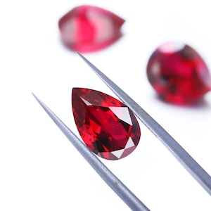 May include: A deep red pear-shaped gemstone held by silver tweezers against a white background.