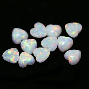 Könnte beinhalten: Eine Sammlung von weißen herzförmigen Opal-Cabochons. Die Opale haben eine milchig weiße Basis mit schillernden Regenbogen-Blitzen.