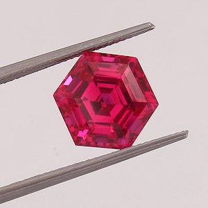 May include: A deep red hexagonal gemstone with a faceted design. The gemstone is held by tweezers.