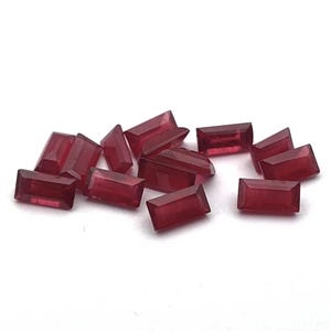 Pode incluir: Um grupo de 12 pedras preciosas retangulares vermelhas, provavelmente rubis, espalhadas sobre uma superfície branca.