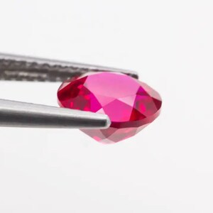 May include: A vibrant red gemstone, likely a ruby, is held by a pair of tweezers. The gemstone is oval-shaped and has a faceted cut, showcasing its brilliance.