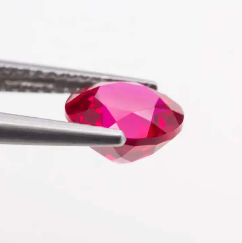 May include: A vibrant red gemstone, likely a ruby, is held by a pair of tweezers. The gemstone is oval-shaped and has a faceted cut, showcasing its brilliance.
