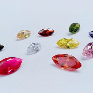 May include: Assortment of marquise-shaped gemstones in various colors, including black, white, red, pink, yellow, green, and purple. The gemstones are arranged on a white surface.