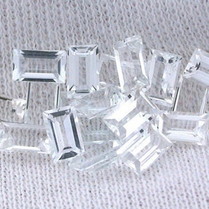 Könnte beinhalten: Eine Gruppe von klaren, rechteckigen, facettierten Edelsteinen, wahrscheinlich Diamanten, die auf einem weißen Stoffhintergrund verstreut sind.