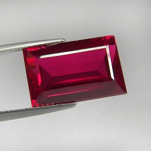 Könnte beinhalten: Ein rechteckiger, facettierter Rubin, der von einer Pinzette gehalten wird. Der Rubin hat eine tiefrote Farbe mit einem rechteckigen Schliff und einer reflektierenden Oberfläche. Der Edelstein ist vor einem schlichten, hellen Hintergrund platziert.