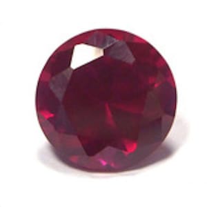 May include: A deep red gemstone with a round, faceted cut. The gemstone is set against a white background.