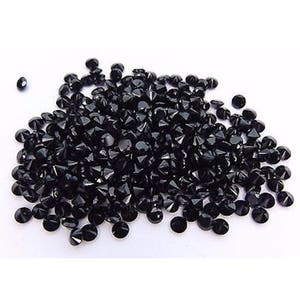 May include: A large pile of black round faceted gemstones. The gemstones are scattered on a white surface.