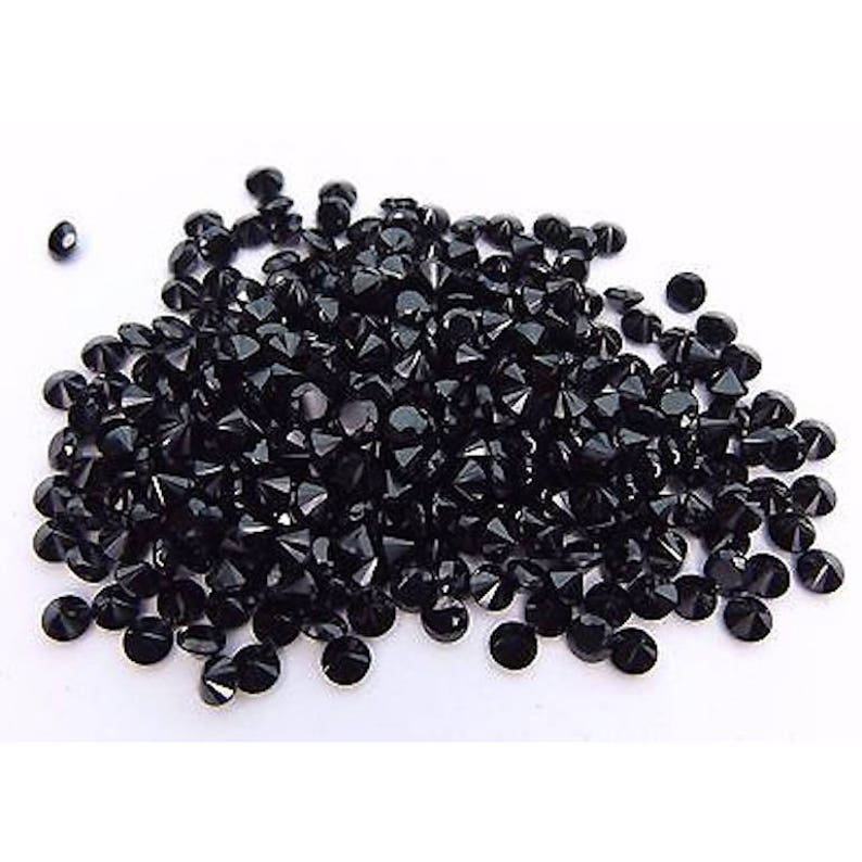 May include: A large pile of black round faceted gemstones. The gemstones are scattered on a white surface.