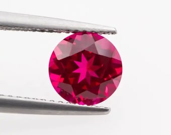 May include: A deep red gemstone with a star-shaped pattern inside, held by tweezers against a white background.