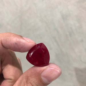 May include: A faceted, triangular-shaped, deep red gemstone. The gem is held between fingers, showcasing its polished surfaces and rich color. The gem's shape is a triangle with rounded corners.
