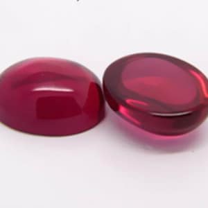 May include: Two polished, cabochon-cut gemstones in a deep red colour. One is a complete oval shape, while the other is a similar oval shape, cut in half. The gems have a smooth, glossy surface, reflecting light.