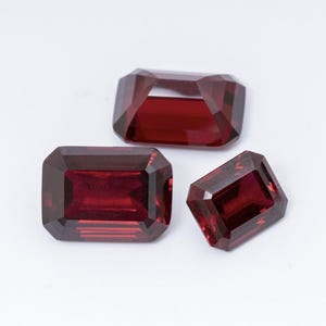 May include: Three faceted, rectangular-cut, deep red gemstones. The gems are displayed against a white background, showcasing their clarity and rich color. The gems are likely garnets, suitable for jewelry.
