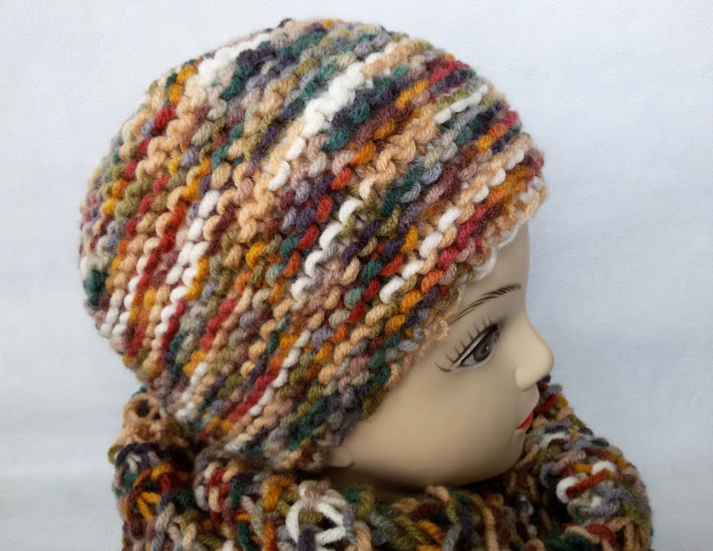 May include: A multi-colored knitted hat and scarf set in shades of brown, orange, green, white, and gray. The hat is a slouchy beanie style and the scarf is a thick, chunky knit.