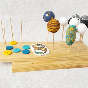 May include: A wooden drying rack with multiple holes for holding clay beads and other crafts. The rack has two levels and is filled with colorful beads, including white, black, blue, yellow, and green.
