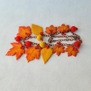 May include: A charm bracelet featuring multiple maple and serrated leaves in shades of yellow, orange, and red. The leaves are interspersed with small red and yellow spherical beads and attached to a copper-toned metal chain with a lobster clasp.