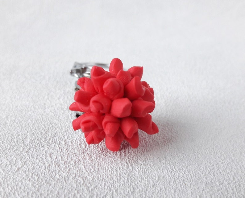Pode incluir: Um vibrante anel floral vermelho. O anel apresenta um design floral tridimensional detalhado em um tom vermelho brilhante. A banda &eacute; prateada, proporcionando um contraste com a flor arrojada. O anel est&aacute; sobre um fundo branco texturizado.