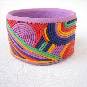 Könnte beinhalten: Ein farbenfrohes, mehrschichtiges Armband aus Ton mit geometrischem Muster. Das Armband besteht aus verschiedenen Tonfarben, darunter Lila, Rot, Gelb, Grün und Blau.