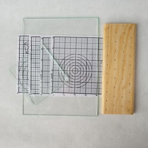 May include: A set of three clear glass rulers with gridded paper underneath. The rulers are marked in inches and have a protractor on one side. A wooden board with holes is on the right side of the image.