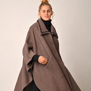 May include: A brown cape with a wide, flowing design. The cape features a collar with black trim and two front pockets. The person in the image is wearing a black turtleneck and black leggings, paired with black boots.