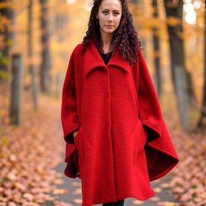 Puede incluir: Una capa de color rojo vibrante hecha de mezcla de lana, con cuello ancho, diseño frontal abierto y bolsillos laterales profundos. La prenda se combina con pantalones vaqueros azul oscuro y zapatillas deportivas de color rosa claro.